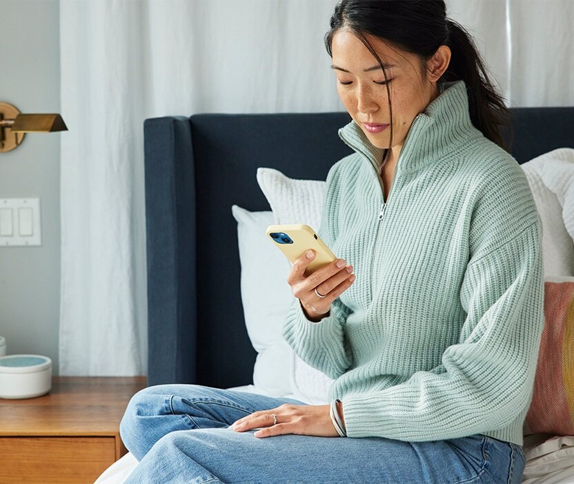 Eine Frau sitzt auf einem Bett, blickt auf ihr Telefon und überlegt, wie sie ein einprägsames Passwort erstellen kann.