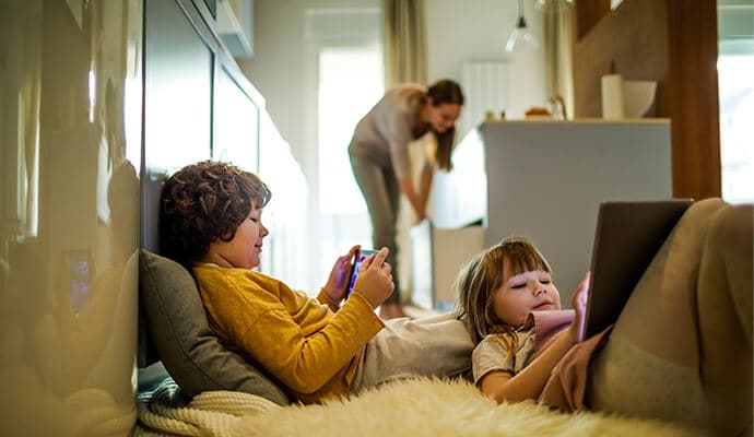 Kinder an Tablets mit Mutter im Hintergrund